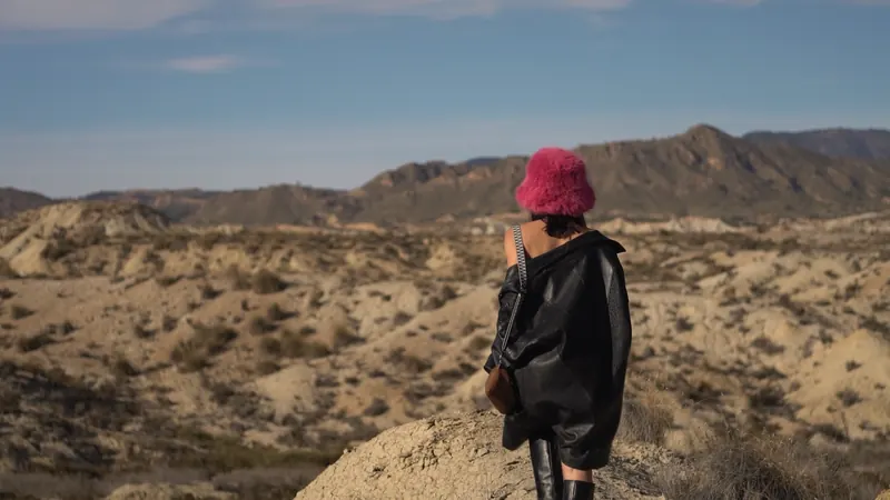 Andrea, con sombrero rosa y chaqueta negra, de pie frente a un paisaje de montaña árido.
