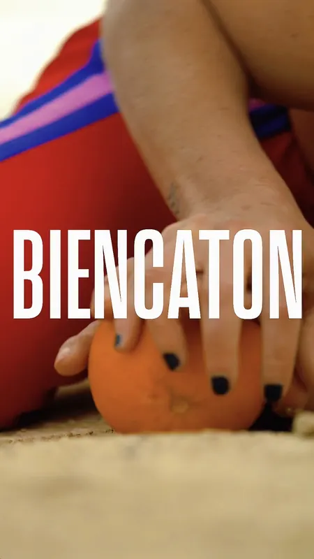 Cartel de Biencaton: una mano con las uñas pintadas sostiene una naranja sobre la arena.