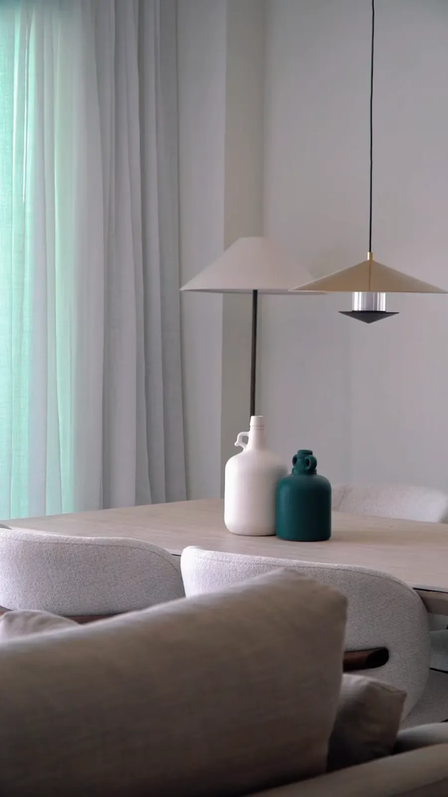 Comedor luminoso con dos lámparas de pantalla cónica, una cortina translúcida y dos botellas de cerámica sobre la mesa.