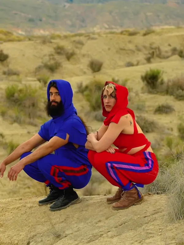 Dos bailarines con ropa roja y azul, agachados en un paisaje árido.