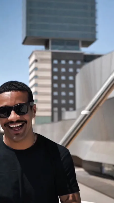 Hombre sonriente con gafas de sol negras frente a una torre de oficinas.