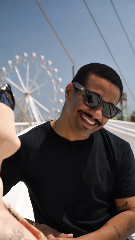 Hombre con gafas de sol y camiseta negra sonríe con una noria al fondo.